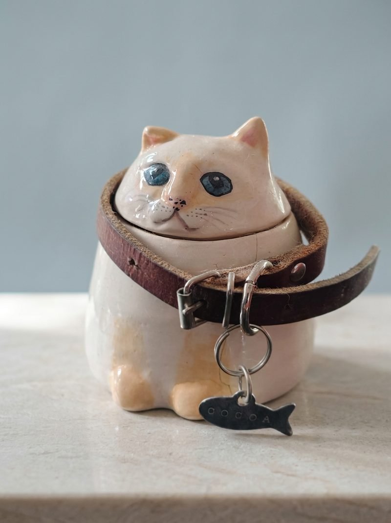 Urna de cerámica de un gato color crema con ojos azules y su collar de piel con placa de pescadito